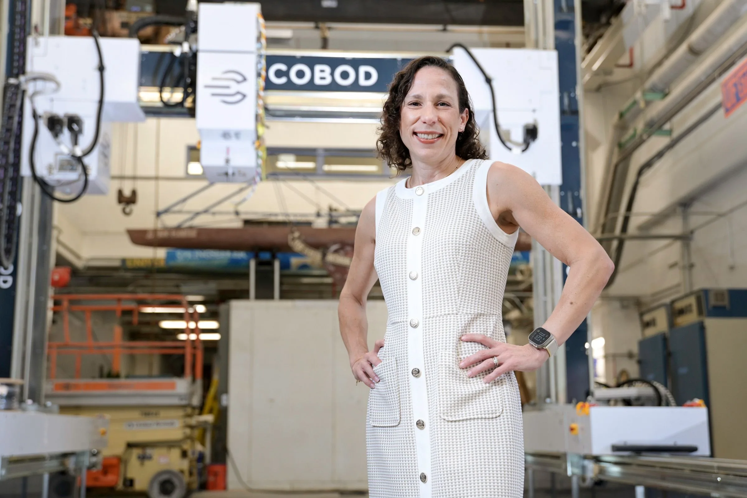 A smiling woman with curly brown hair stands with hands on hips in a large industrial workshop with machinery and a lift.