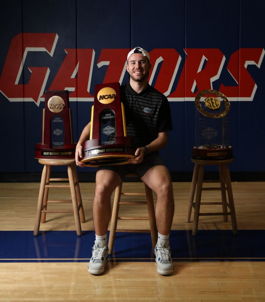 Corey Reynolds sitting in the basketball gym holding a championship trophy, with two additional trophies displayed beside him.