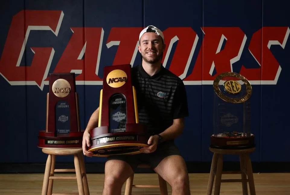 Corey Reynolds sitting in the basketball gym holding a championship trophy, with two additional trophies displayed beside him.
